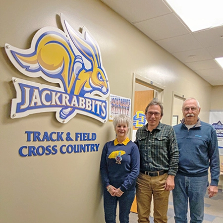 Three people standing in front of a SDSU track and field/cross country sign
