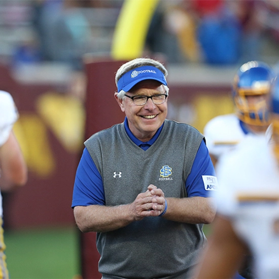 Former head coach John Stiegelmeier smiling on the football field in his coaching gear.