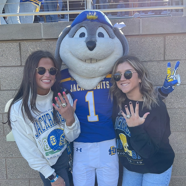 payton burckhard stands with another girl and jack the jackrabbit wearing their summit league championship rings