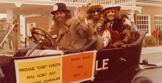 The first Dirty Lil and Students' Association president, Barb Strandell, and Weary Wil, Jack Soukup, and riding in the Bummobile which is being driven by Michael 'Chief' Eveleth, 1976 Hobo Day committee chairman and Grand Pooba