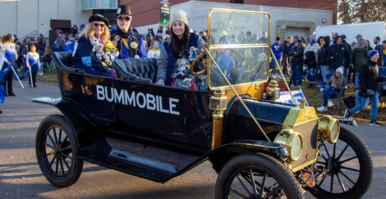 Bummobile drives in the hobo day parade with the South Dakota Art Museum in the background and the people inside of it smiling and waving