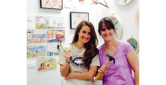 Grace and her mom stand in an art exhibit with artwork behind them and hammers in their hands
