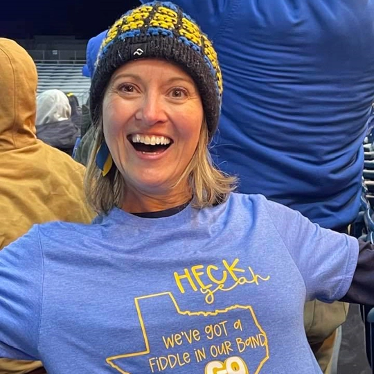 Julie Mooney, smiling ear to ear, as she reveals her shirt that says "Heck yeah, we've got a fiddle in our band. Go Jacks!"