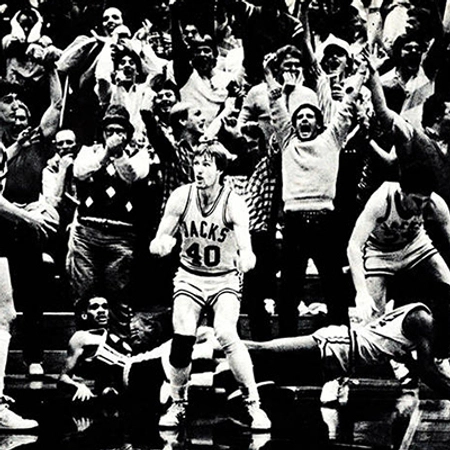 A black and white photo from Frost arena. A jacks basketball player looks on to see if the shot was make while the opposing played are laying on the ground behind him. The crowd has their hands up cheering happily.