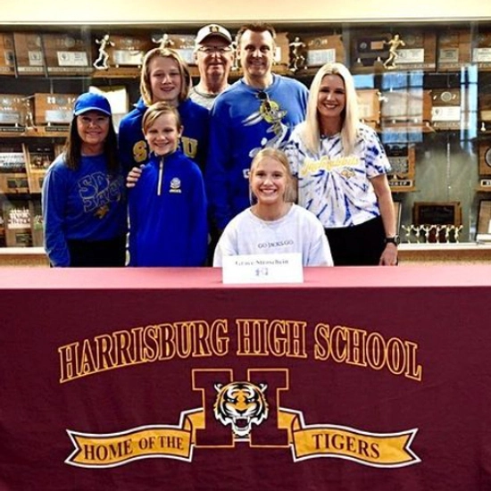 grace stroschein smiling and signing a piece of paper with her family behind her