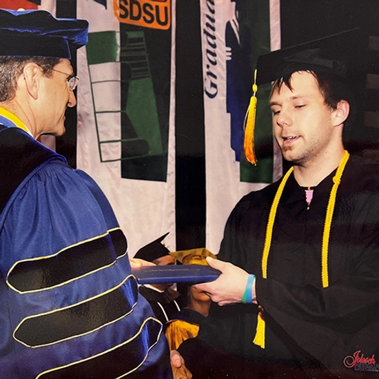 A young man wearing his cap and gown receiving his diploma from a dean of the college