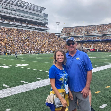 Julie and Marc Mooney smile standing on a football field with yellow and black crowd filled stands in the background