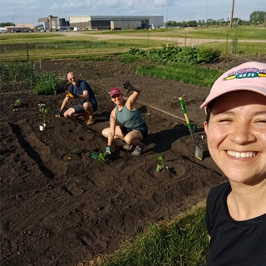 Anne Karabon waving at the camera while planting a garden at the Brookings community gardens with her partner, Matt, and GA, Laura.