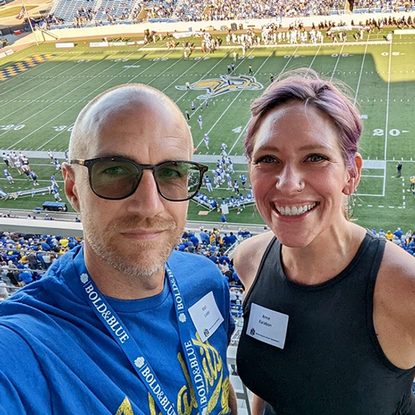 Anne Karabon and her partner, Matt, smiling at the camera while standing in one of the suites overlooking the SDSU football field.