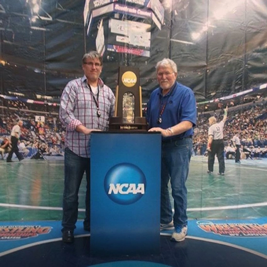 Frank and his son, Steve, smiling for the camera at a NCAA wrestling tournament, posing with a large trophy.