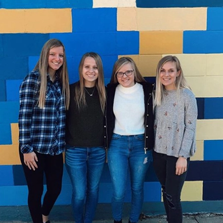 group of 4 women standing with a blue and yellow painted mural behind them