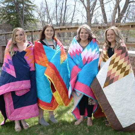 4 female students during their honoring ceremony, wrapped in colorful star quilts