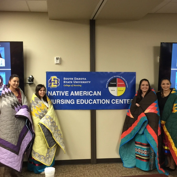 4 female students during their honoring ceremony, wrapped in colorful star quilts standing next to a banner that reads Native American Nursing Education Center (NANEC)