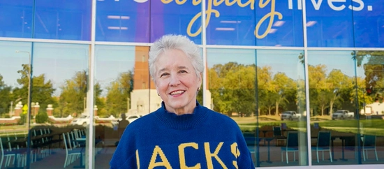A lady wearing a jacks sweatshirt standing in front of the alumni & foundation building smiling