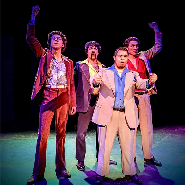 Robert stands with three other young men in 70-inspired suits with stage makeup on in a theatre performance.