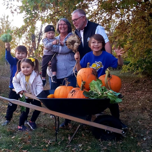 john and laurie steigelmeier stand outdoors with their grandkids and a wheelbarrow of pumpkins