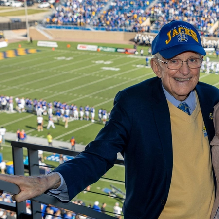 Jim woster smiles wearing a blue jacks hat with the dana j dykhouse stadium field in the background