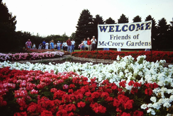 A lush garden with vibrant red, pink, and white flowers in bloom. A sign reads "Welcome Friends of McCrory Gardens." People stroll in the background.