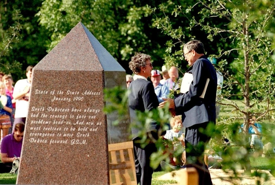 A man and a woman stand beside a stone monument with inscriptions in McCrory Garden park.