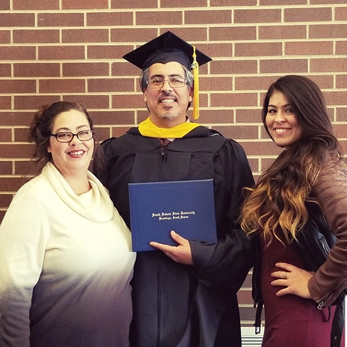 Freddy Sr. holds an SDSU diploma while wearing his commencement attire, standing next to Debra and Sam Moran.