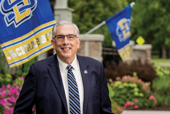 President Barry H. Dunn smiles outside the University Home with beautiful landscaping and SDSU flags in the background.
