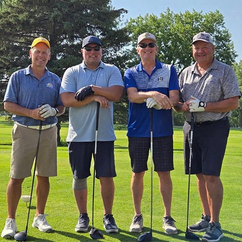 Four men stand on a sunny golf course, smiling, each holding a golf club.