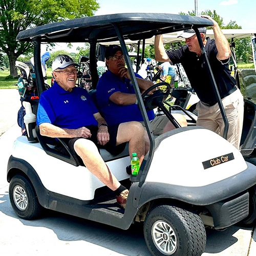 Three men are gathered around a golf cart, two sitting and one standing.