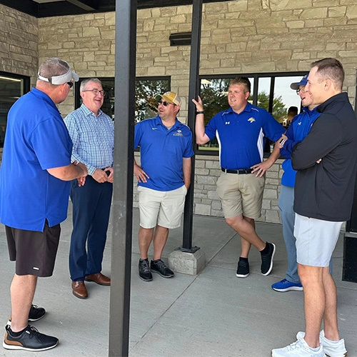A group of six men casually chat outside a stone building.