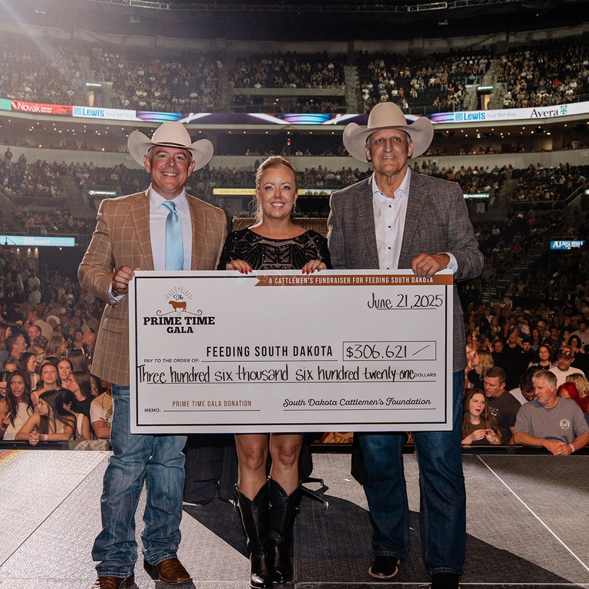 Three people standing together on a stage holding up a large check with an arena full of people in the background.