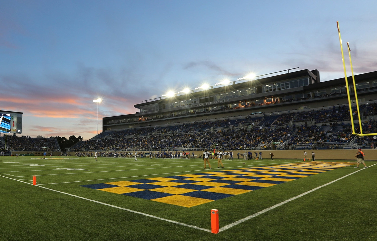 A packed football stadium at dusk, with players on the field, a giant midfield logo, bright lights, and yellow goalposts.
