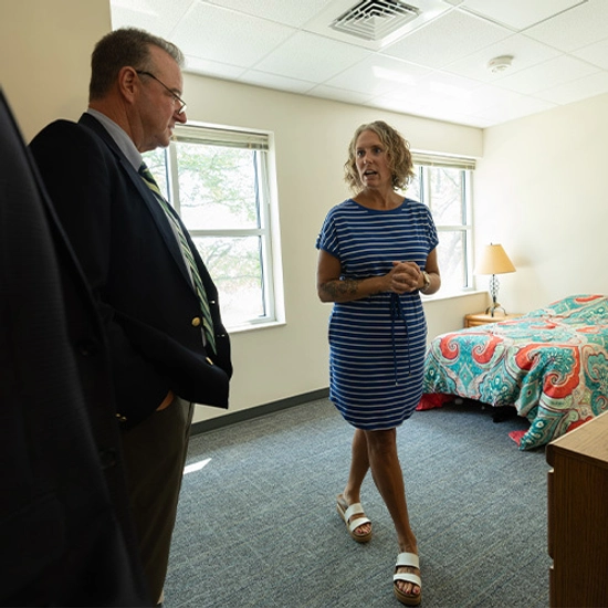 A woman in a striped dress speaks with a suited man in a bright bedroom with patterned bedding, conveying a calm, professional conversation.
