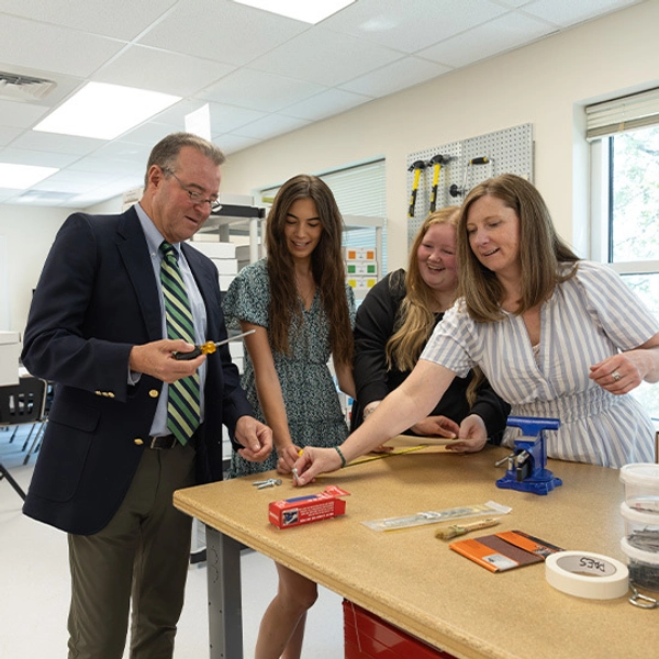 A suited man and three women gather at a classroom workbench, examining a small project with tools and smiling in a collaborative mood.
