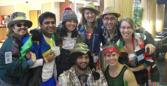 A cheerful group of eight friends pose closely together indoors, wearing quirky hats, scarves, and colorful costumes; the mood is playful and festive.