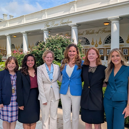 Six women smile for a group photo outside the white-columned Rose Garden Hall of Fame, with flowering greenery and a bright, welcoming feel.