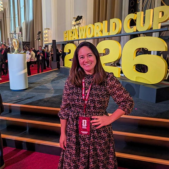 Susanna Marking poses beside oversized gold “FIFA World Cup 2026” letters at an indoor event, with a trophy display and guests in the background.