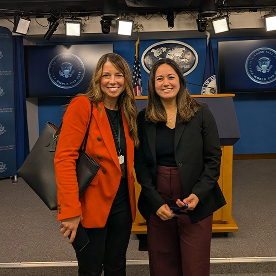 Two smiling women pose in front of a White House press briefing podium and presidential seals, beneath studio lights.