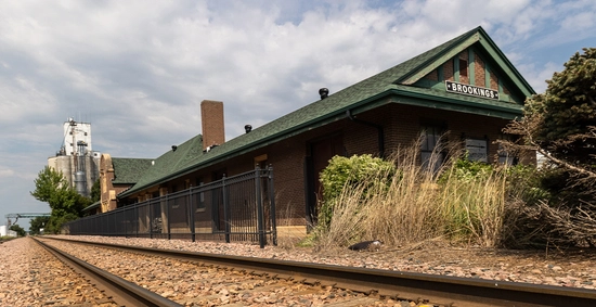 A weathered rural train station with a green gabled roof sits beside empty railroad tracks, framed by dry grasses beneath a cloudy sky.
