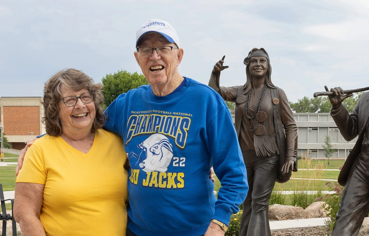 A smiling couple stands beside the bronze statues of Dirty Lil and Weary Wil on campus at South Dakota State University.