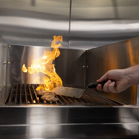 A hand flipping burger patties on a grill with a flame rising from the grill.