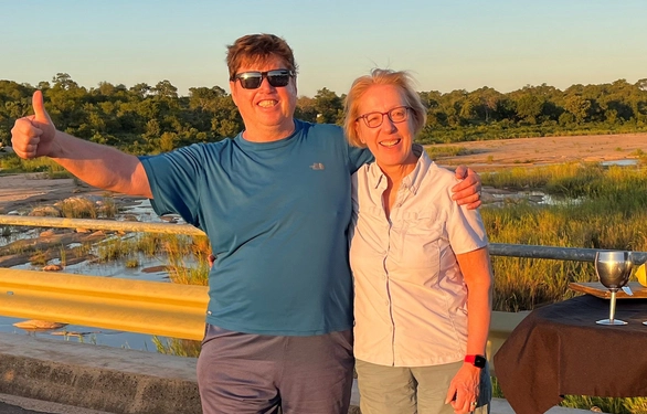 A smiling man in sunglasses gives a thumbs-up beside a smiling woman on a sunlit boardwalk.