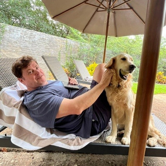 A relaxed man lounges beneath a patio umbrella, smiling as he pets a golden retriever sitting beside him outdoors.