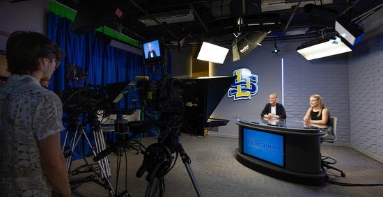 Two students sit at the desk in the studio in Yeager Hall while more students stand behind the camera and film them.
