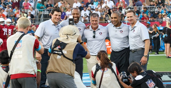 Former NFL players and staff pose for a cheerful field-side photo as photographers capture them, with a packed stadium crowd in the background.