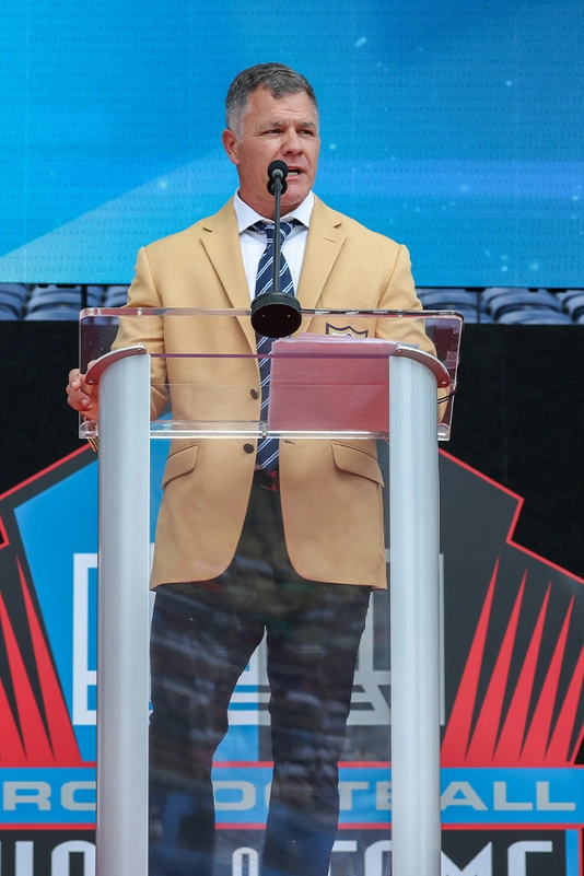 Adam Vinatieri wears a tan blazer speaking at the Hall of Fame podium beside his football bust and framed player portrait.