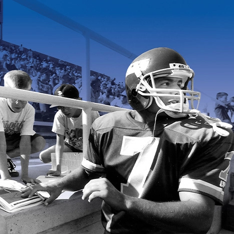 A black and white photo of Adam Vinatieri signing a program on the sideline of the SDSU football stadium.