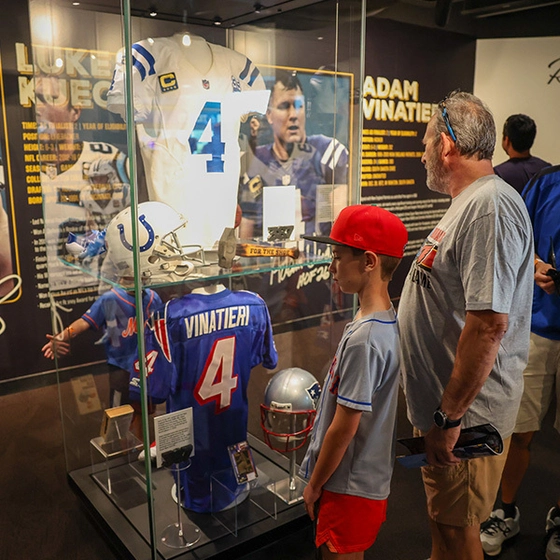 Visitors view an Adam Vinatieri football exhibit with number 4 jerseys, helmets, and photos in a glass case, creating an informative museum atmosphere.