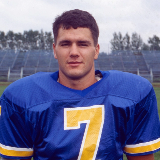 A young Adam Vinatieri in a blue jersey with a large yellow number 7 standing on a football field.