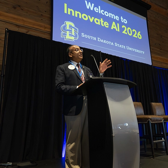 A speaker in formal attire gives a presentation at a podium on a stage, with a large screen behind him that reads "Welcome to Innovate AI 2026."