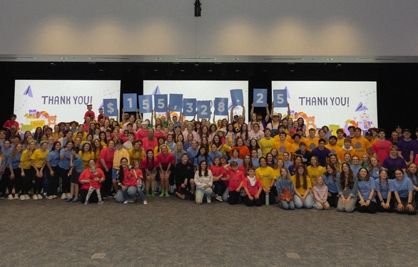 A large group of people in colorful shirts holding signs with numbers, posed in a spacious hall with two screens displaying "Thank You!"
