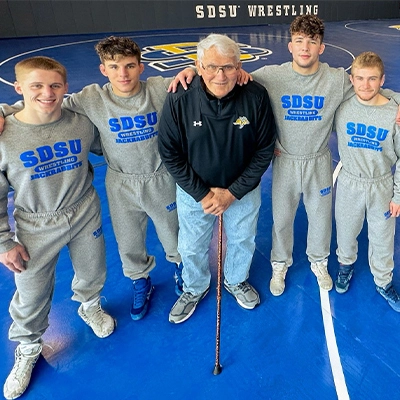 Frank Kurtenbach stands on a wrestling mat with 4 SDSU wrestlers next to him.